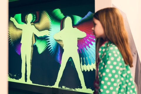 Ein Mädchen steht seitlich im Bild, im Hintergrund sind ihre Umrisse auf einer Leinwand mit vielen verschiedenen Farben umrandet.